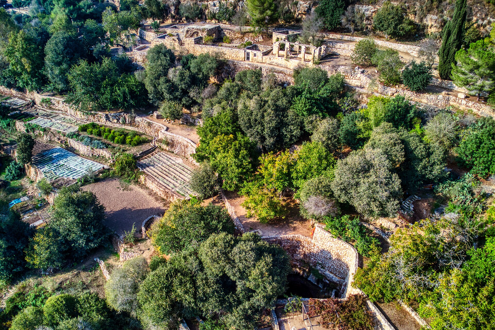 Aerial view of Sataf with stone-walled farming terraces and vegetable plots, restored village ruins along a path above and a curved spring pool wall below
