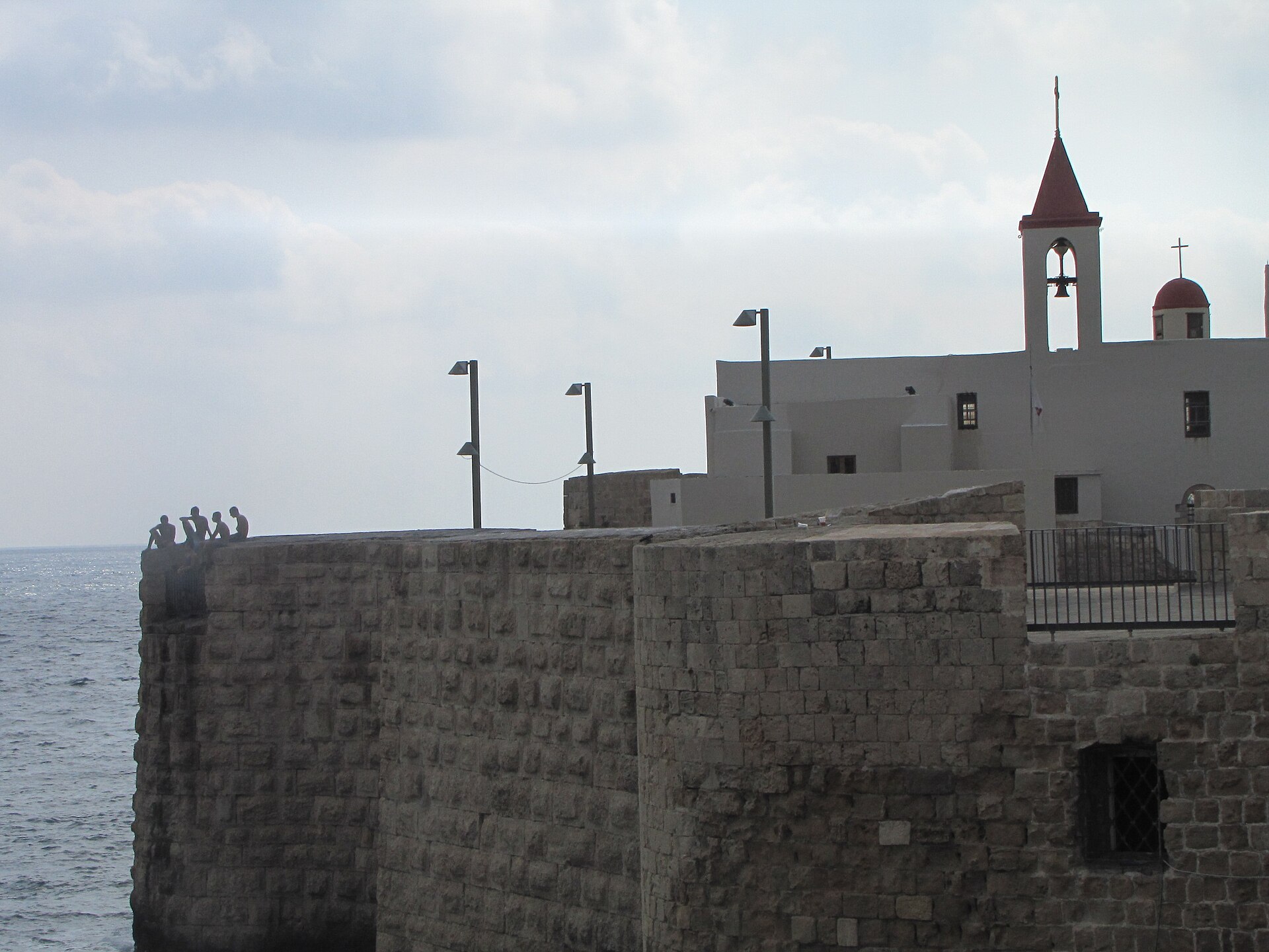 Teenage boys sitting on top of Akko's stone sea wall above the Mediterranean, with a church bell tower behind