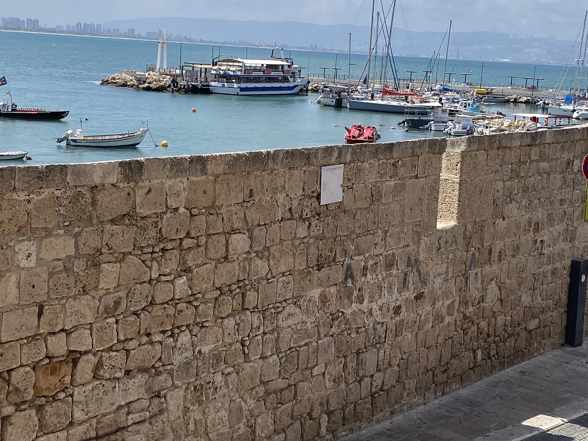 Akko's old stone sea wall overlooking the fishing harbour, with moored boats and a tour boat, and Haifa and Mount Carmel across the bay