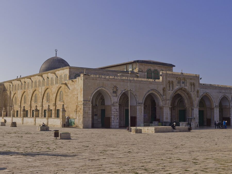 The Al-Aqsa Mosque's seven-arched stone portico and grey lead dome seen across the open paved esplanade of the Temple Mount / Haram al-Sharif, with a few visitors