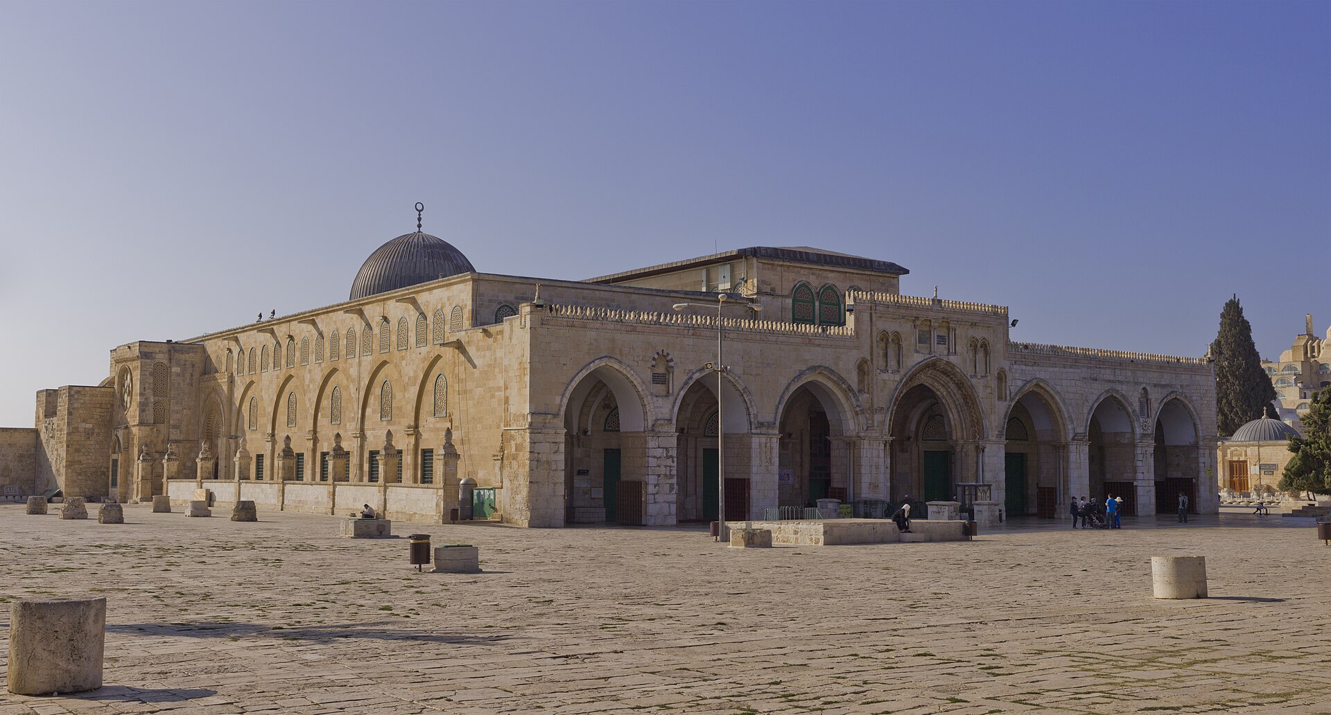 The Al-Aqsa Mosque's seven-arched stone portico and grey lead dome seen across the open paved esplanade of the Temple Mount / Haram al-Sharif, with a few visitors