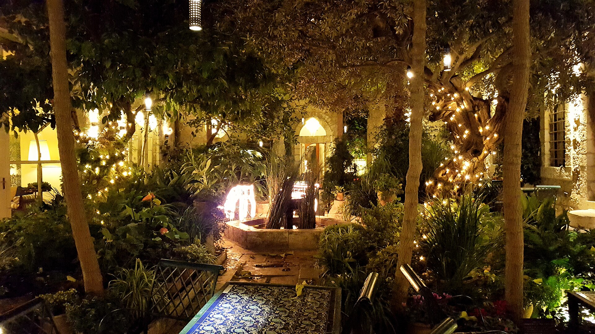 The American Colony Hotel's inner courtyard at night, with fairy lights wrapped around an old olive tree, a stone fountain, lush plants and a tiled table