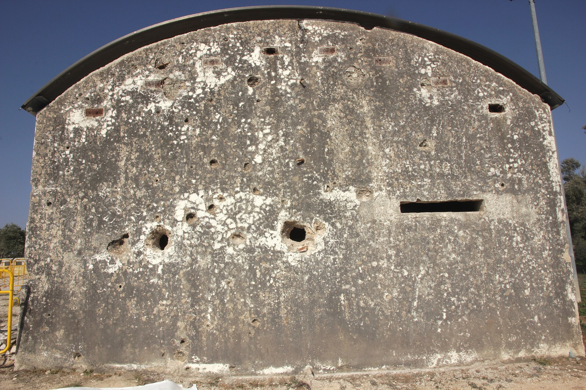 The pale concrete end wall of a building at the top of Ammunition Hill, pitted with bullet and shell holes, under a curved metal roof