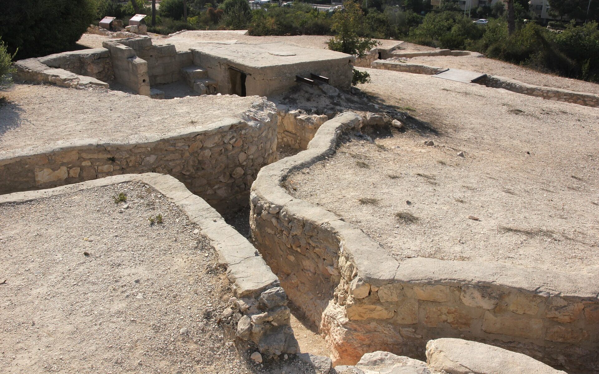 Zigzagging stone-lined trenches leading to a concrete bunker on Ammunition Hill in Jerusalem, with information signs and trees behind