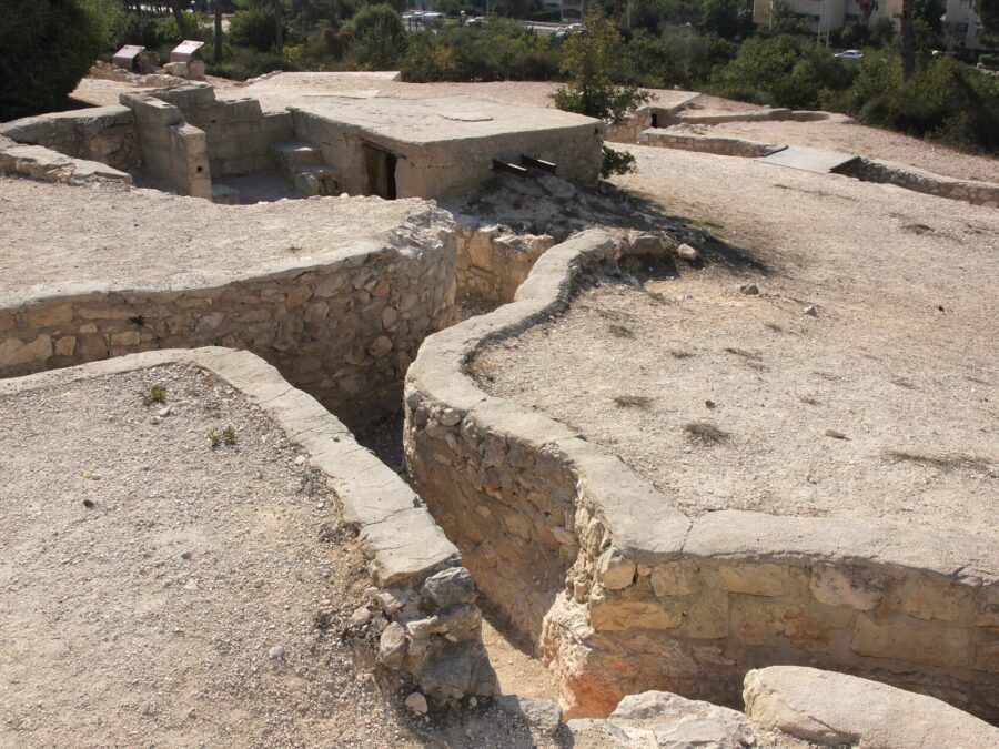 Zigzagging stone-lined trenches leading to a concrete bunker on Ammunition Hill in Jerusalem, with information signs and trees behind