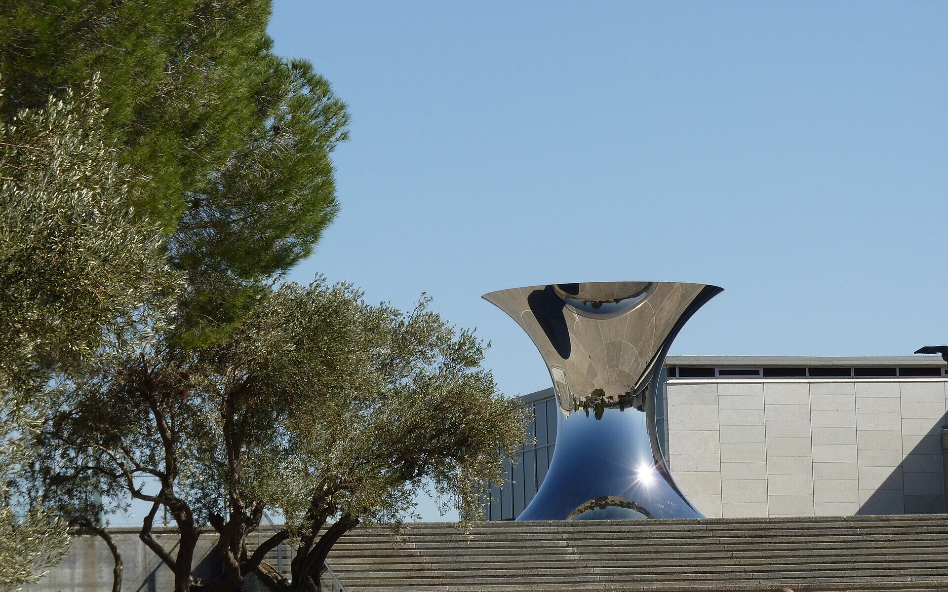Anish Kapoor's polished steel hourglass sculpture reflecting the sky above broad stone steps at the Israel Museum, with olive and pine trees and a stone-clad pavilion