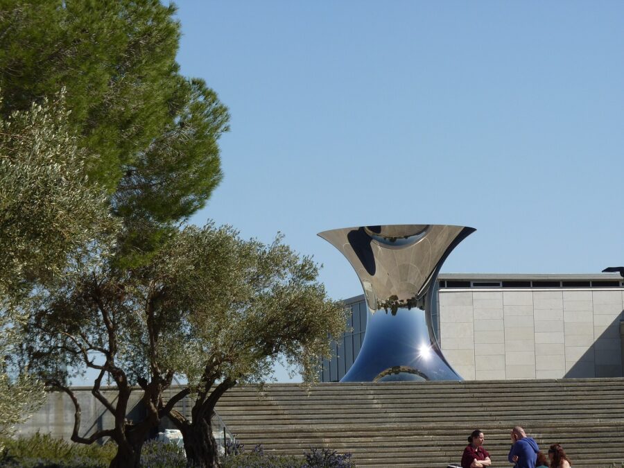 Anish Kapoor's polished steel hourglass sculpture reflecting the sky above broad stone steps at the Israel Museum, with olive and pine trees and a stone-clad pavilion