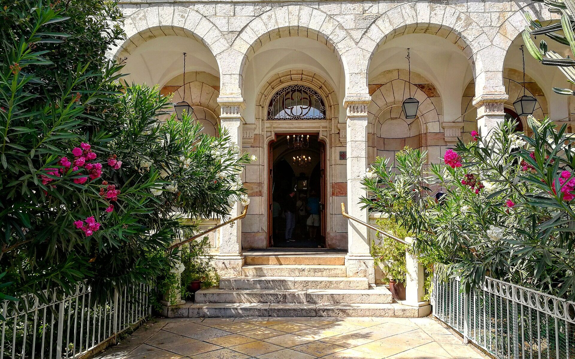 Arcaded stone entrance of the Austrian Hospice in Jerusalem's Old City, with steps up to an open door framed by pink and white oleanders and hanging lanterns
