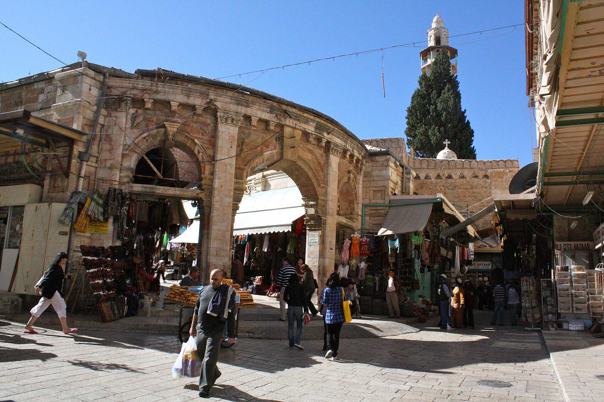 The arched Souq Aftimos gateway in the Muristan, Jerusalem's Christian Quarter, with clothing and leather shops, a sesame bread cart and the Mosque of Omar minaret