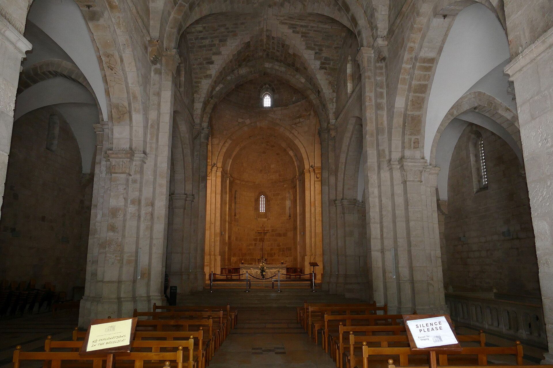 Bare stone nave of the Crusader Church of St Anne in Jerusalem, with pointed arches, massive piers and wooden pews leading to a softly lit apse and altar