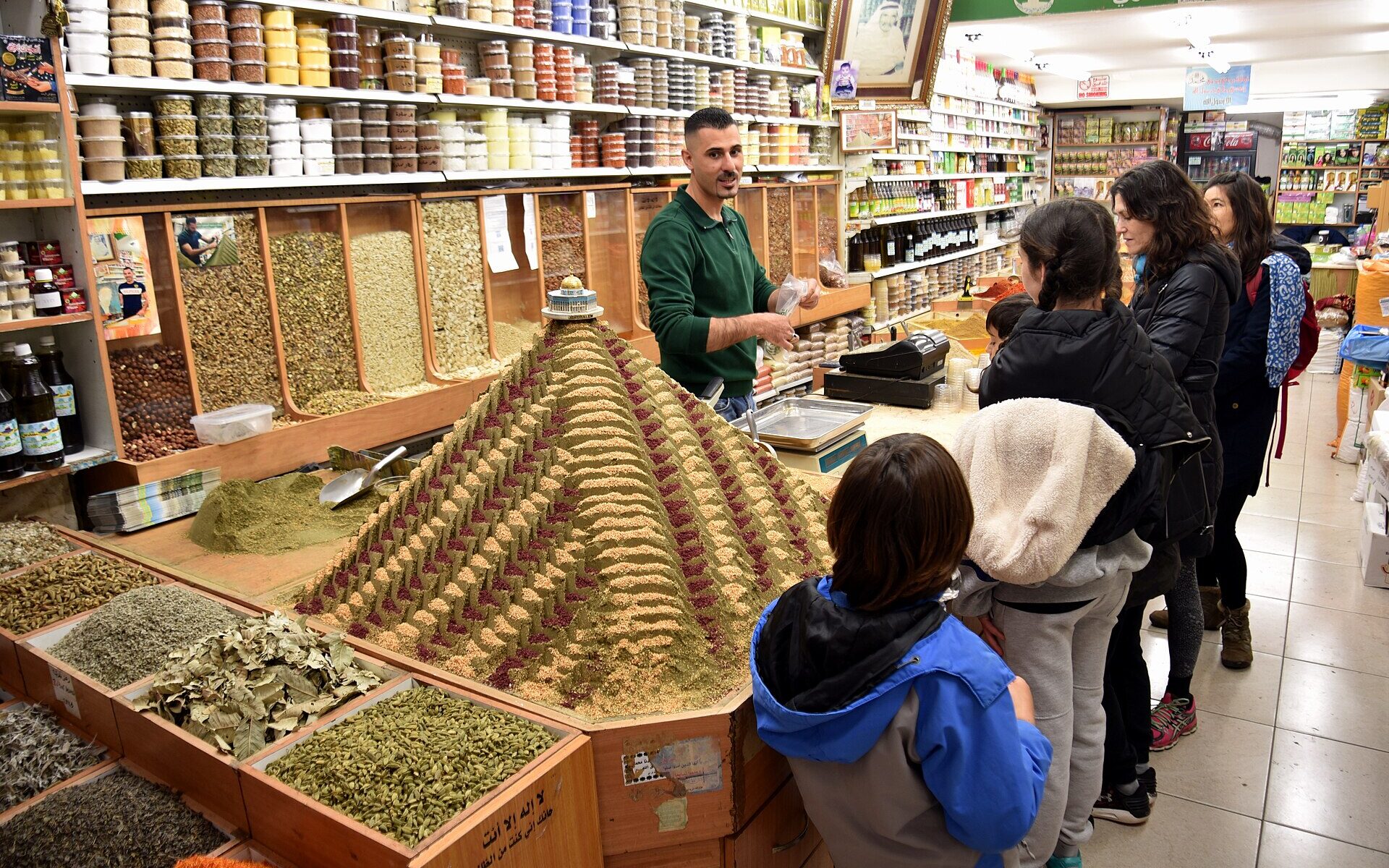 Behind a spice pyramid topped with a Dome of the Rock model, a vendor serves a family in an Old City spice shop lined with bins of herbs
