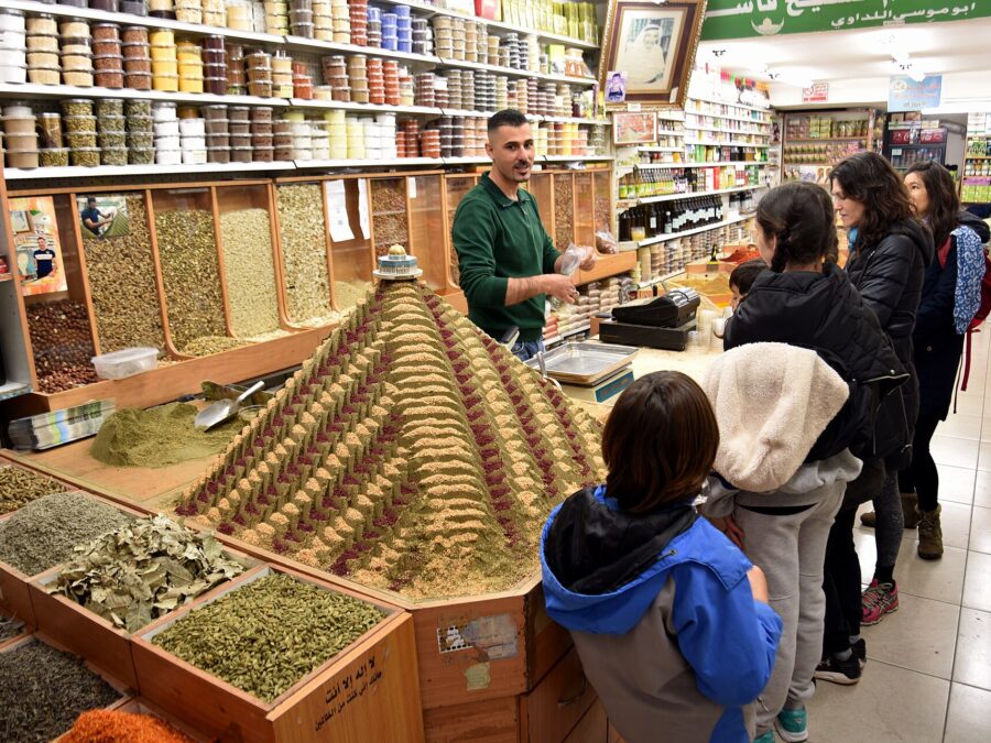 Behind a spice pyramid topped with a Dome of the Rock model, a vendor serves a family in an Old City spice shop lined with bins of herbs