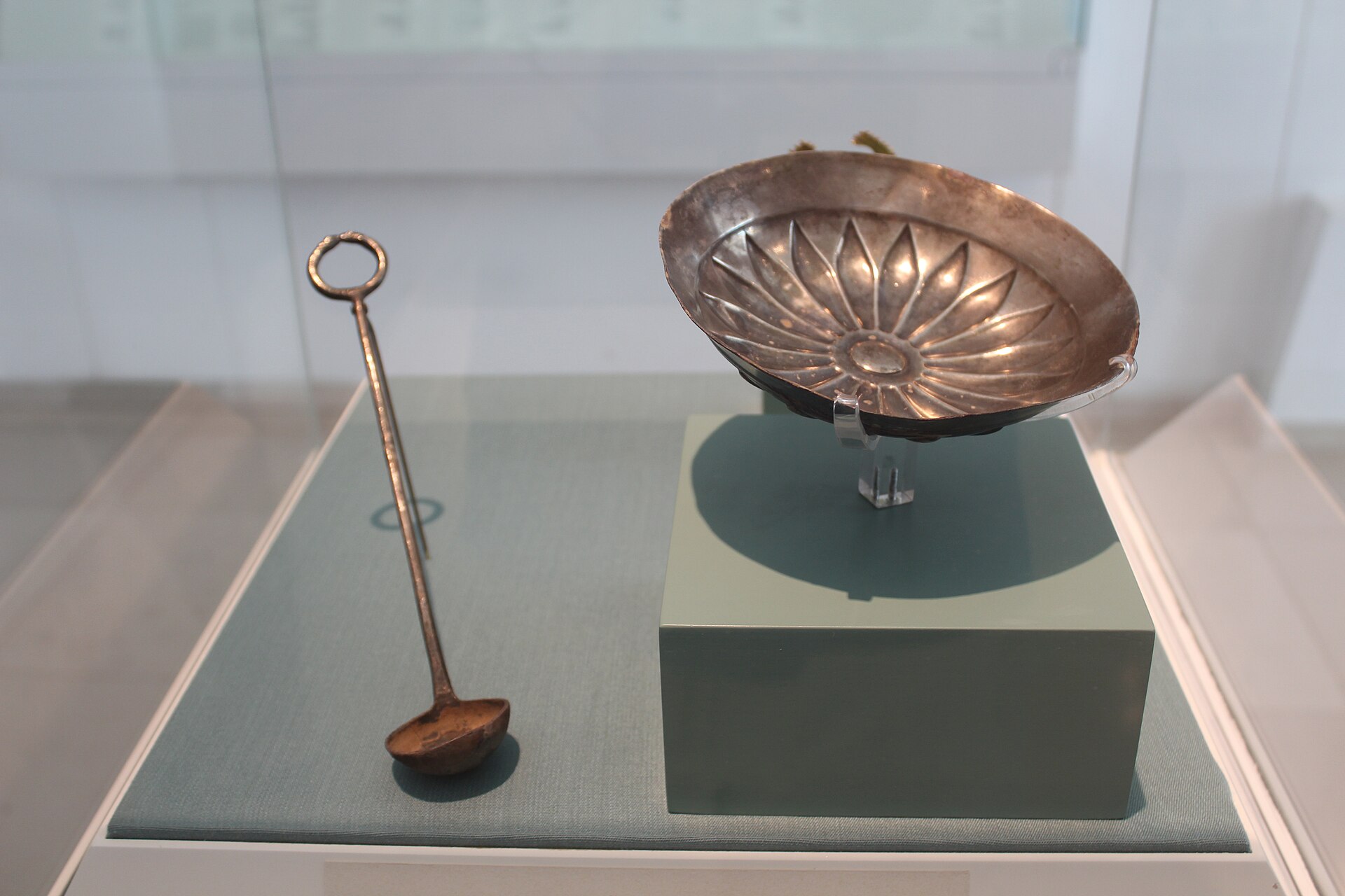 An ancient silver bowl with a fluted, petal-shaped interior and a long-handled silver ladle displayed in a glass case at the Bible Lands Museum in Jerusalem