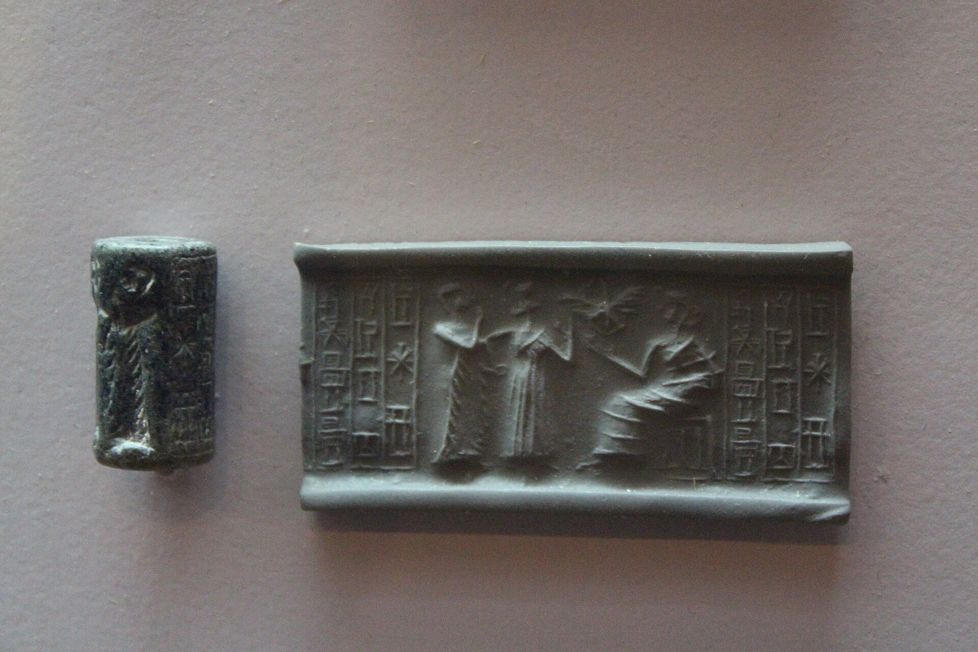 A dark stone cylinder seal beside the clay impression it makes, showing robed figures before a seated deity and columns of cuneiform text, at the Bible Lands Museum in Jerusalem
