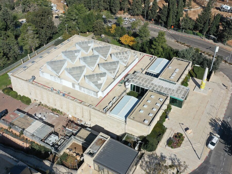Aerial view of the Bible Lands Museum in Jerusalem, a square stone building with a roof of white pyramid-shaped skylights, a glass entrance wing and a plaza with a tall column
