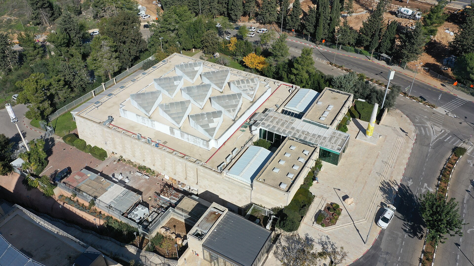 Aerial view of the Bible Lands Museum in Jerusalem, a square stone building with a roof of white pyramid-shaped skylights, a glass entrance wing and a plaza with a tall column