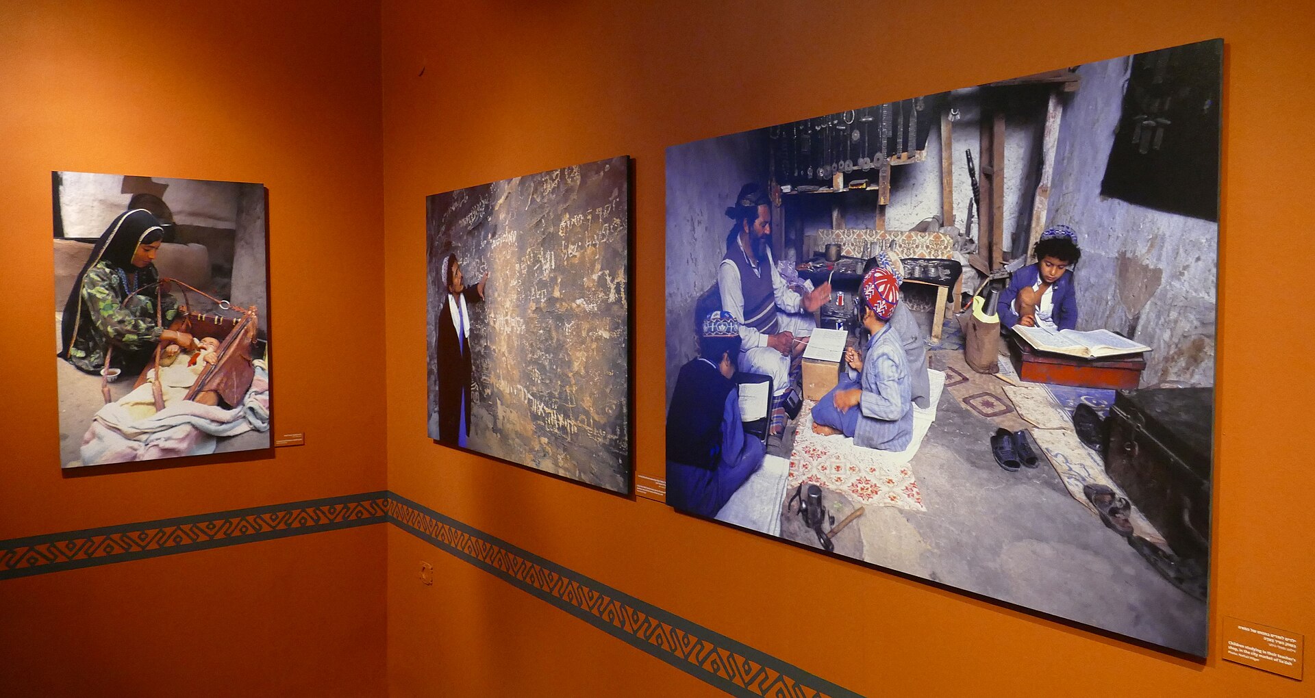 Large colour photographs of Jewish life in Yemen, including children studying with a teacher and a mother with a baby in a cradle, hung on orange walls in a temporary exhibition at the Bible Lands Museum