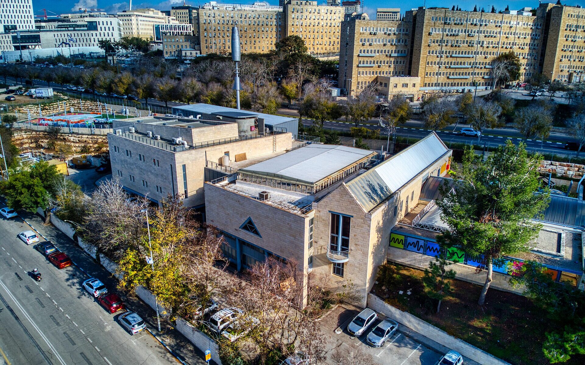 Aerial view of the Bloomfield Science Museum in Jerusalem, with pale stone buildings, a glass-roofed hall and brightly coloured window panels, and government office blocks behind