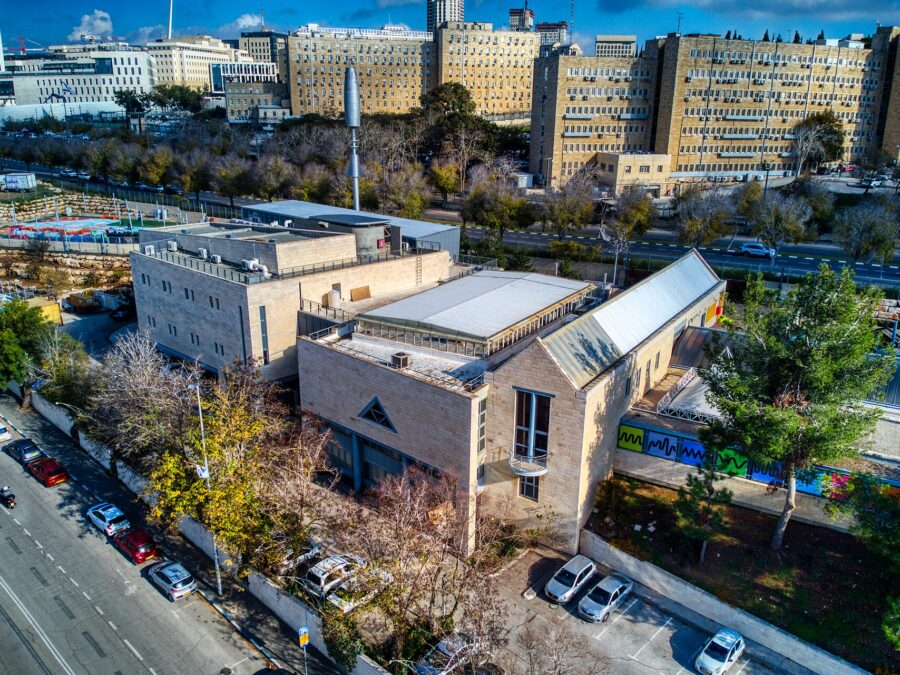 Aerial view of the Bloomfield Science Museum in Jerusalem, with pale stone buildings, a glass-roofed hall and brightly coloured window panels, and government office blocks behind