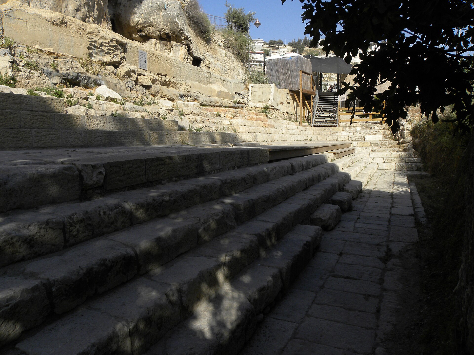 Broad flights of ancient stone steps along the excavated Pool of Siloam, with a wooden boardwalk over the upper tier and a shaded platform beyond