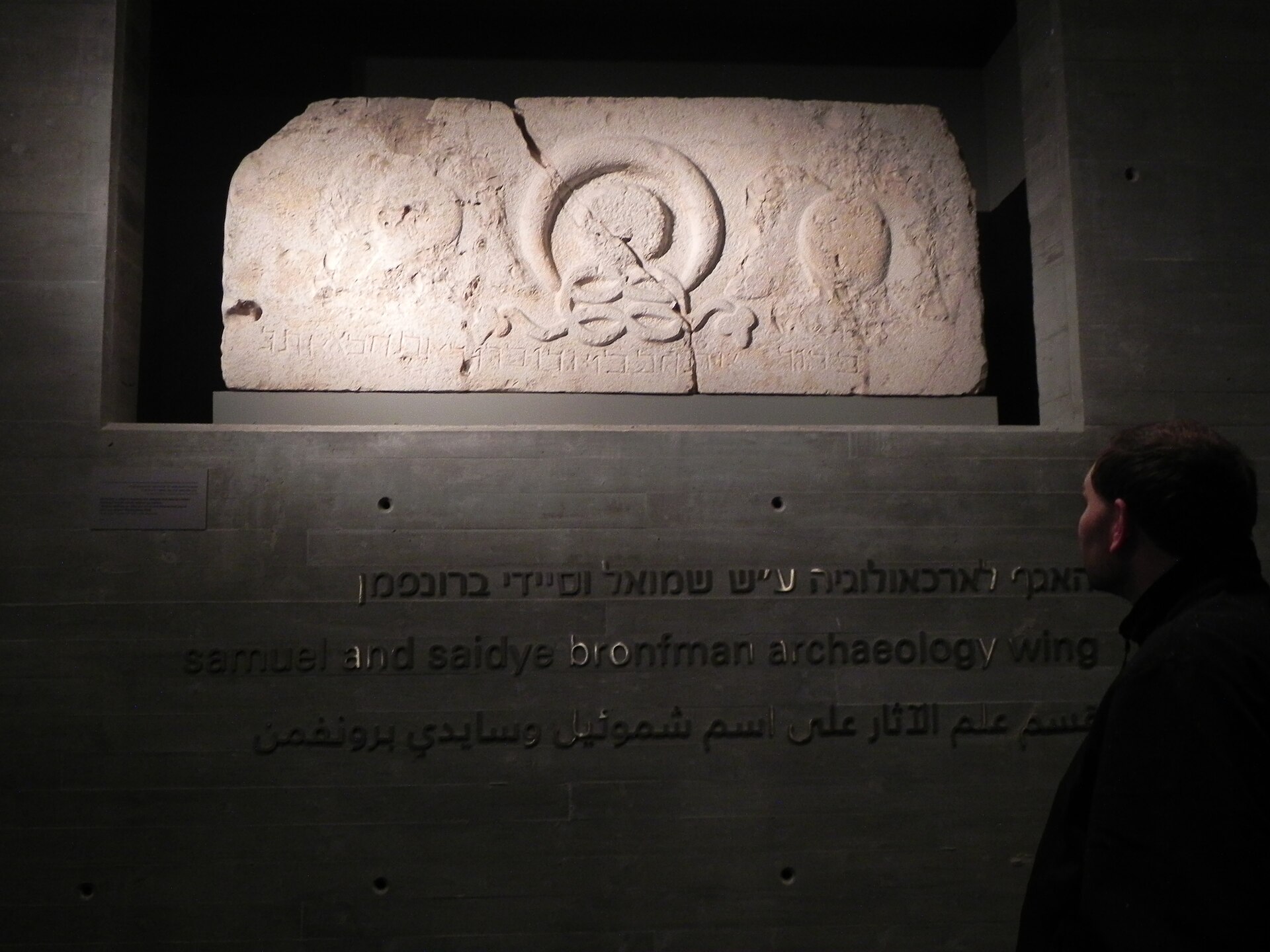 A carved stone lintel with a wreath and an inscription displayed above the name of the Samuel and Saidye Bronfman Archaeology Wing at the Israel Museum, with a visitor looking on
