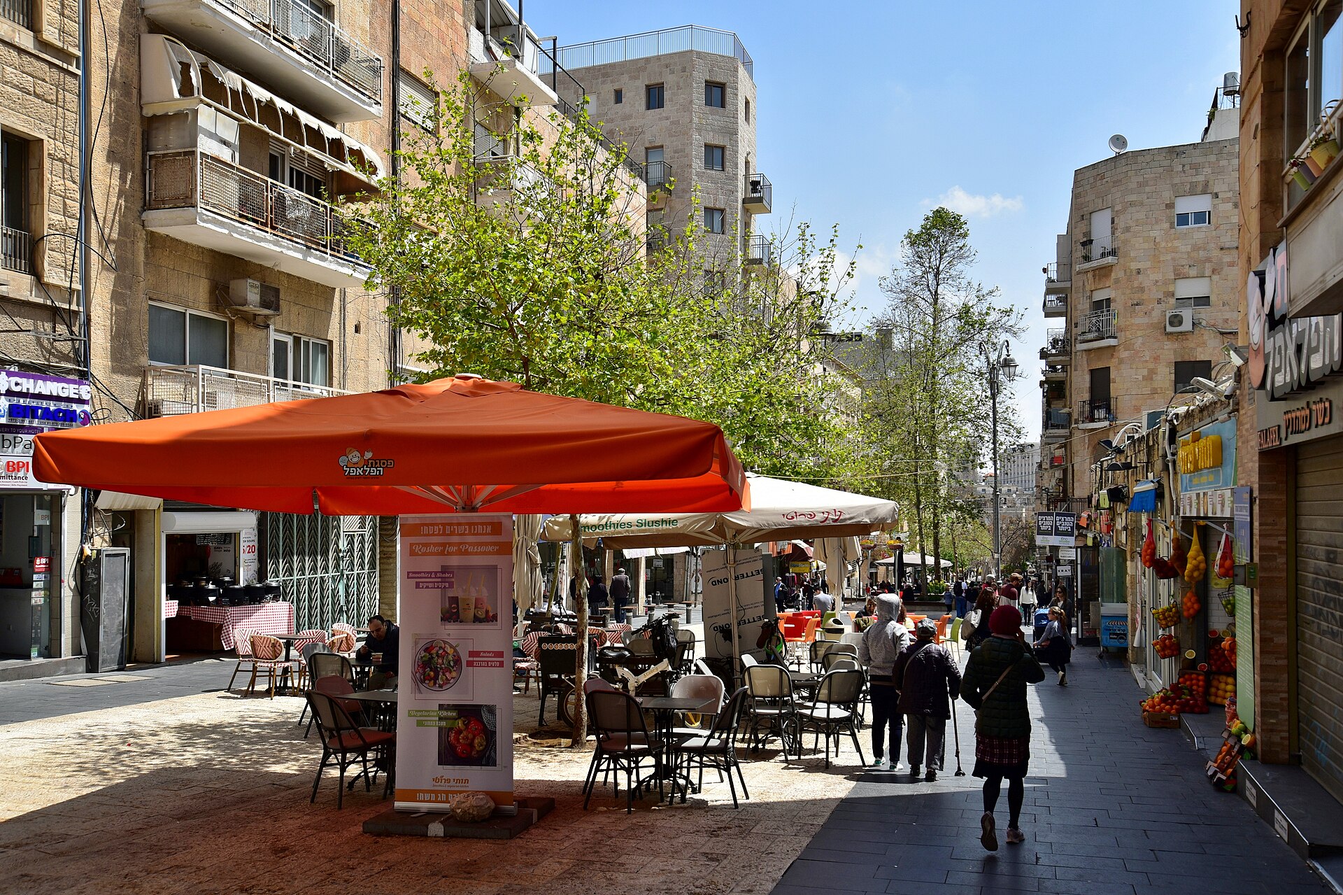 Café tables under orange parasols, a falafel shop and a fruit-juice stall on the Ben Yehuda Street pedestrian mall in Jerusalem, with stone apartment blocks and pedestrians