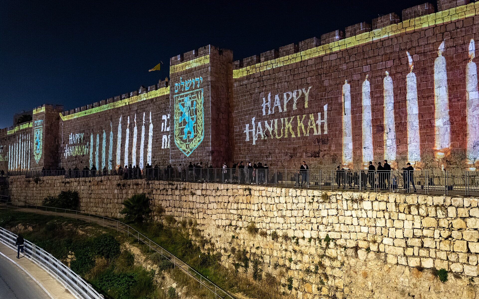 Candles, the Jerusalem city emblem and 'Happy Hanukkah' projected in light onto the Old City walls at night, with people walking along the path below