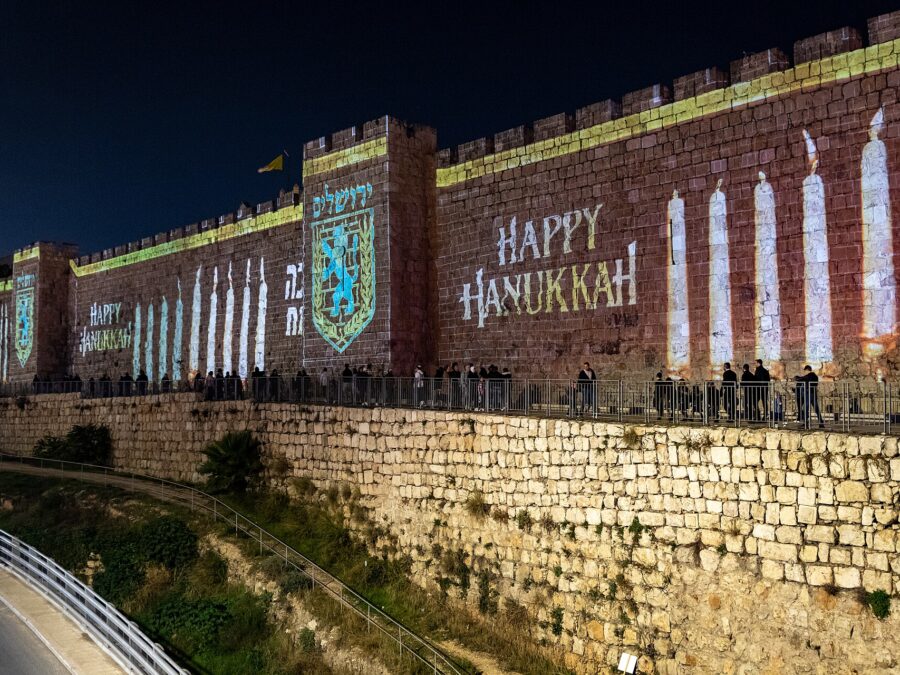 Candles, the Jerusalem city emblem and 'Happy Hanukkah' projected in light onto the Old City walls at night, with people walking along the path below