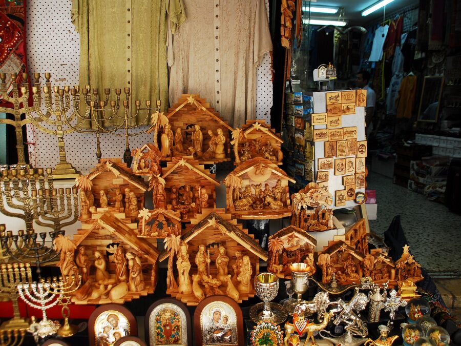 Carved olive-wood nativity scenes stacked in an Old City shop display in Jerusalem, beside brass menorahs, silver-framed icons, kiddush cups and olive-wood magnets