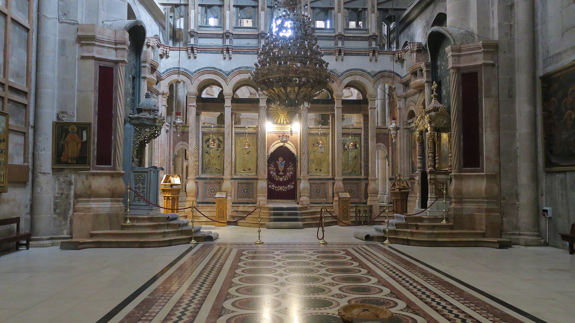 The Catholicon inside the Church of the Holy Sepulchre, with an inlaid marble floor, a large chandelier, two carved thrones and a gilded iconostasis hung with icons