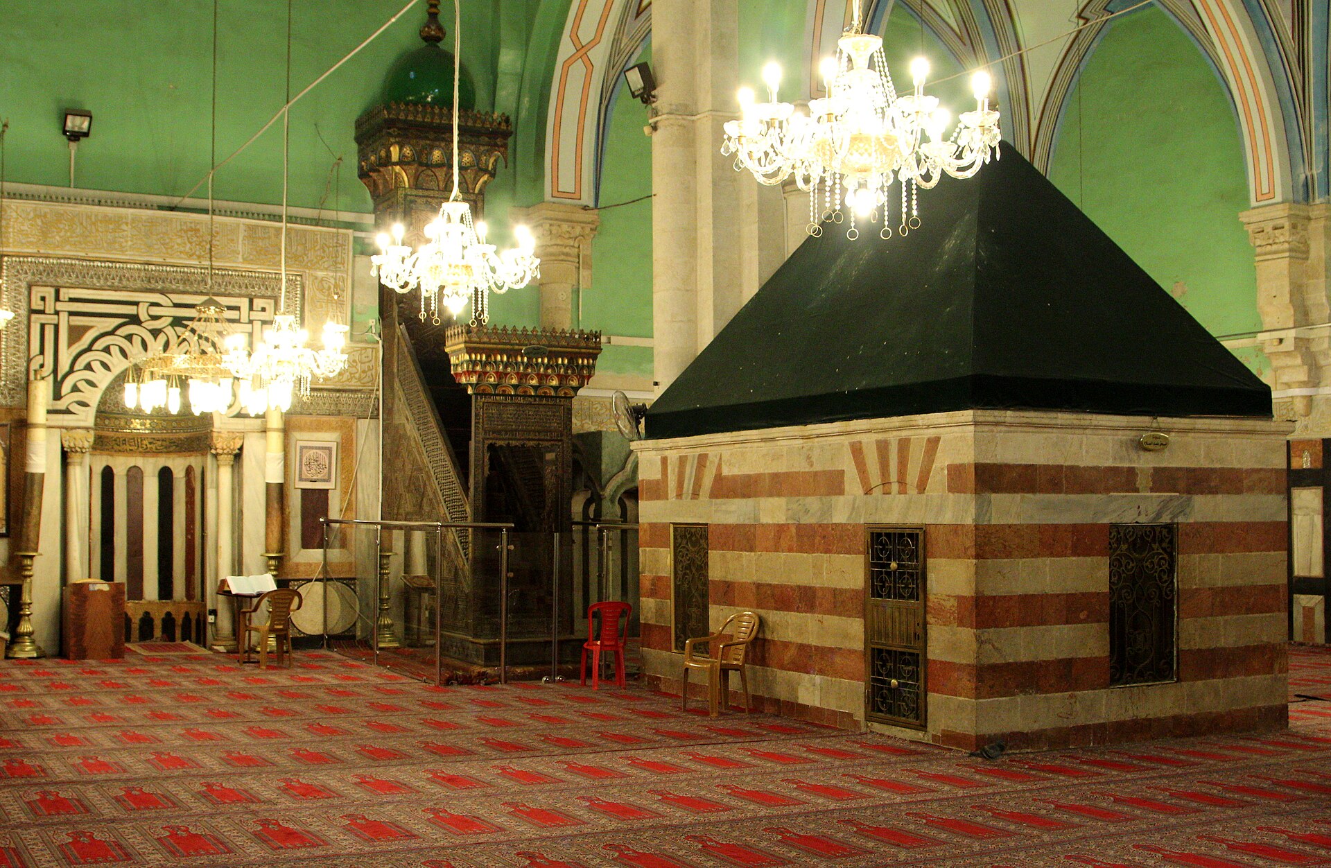 The cenotaph of Isaac inside the Ibrahimi Mosque at the Cave of the Patriarchs in Hebron, a striped marble shrine with a dark pyramid roof beside a carved wooden pulpit