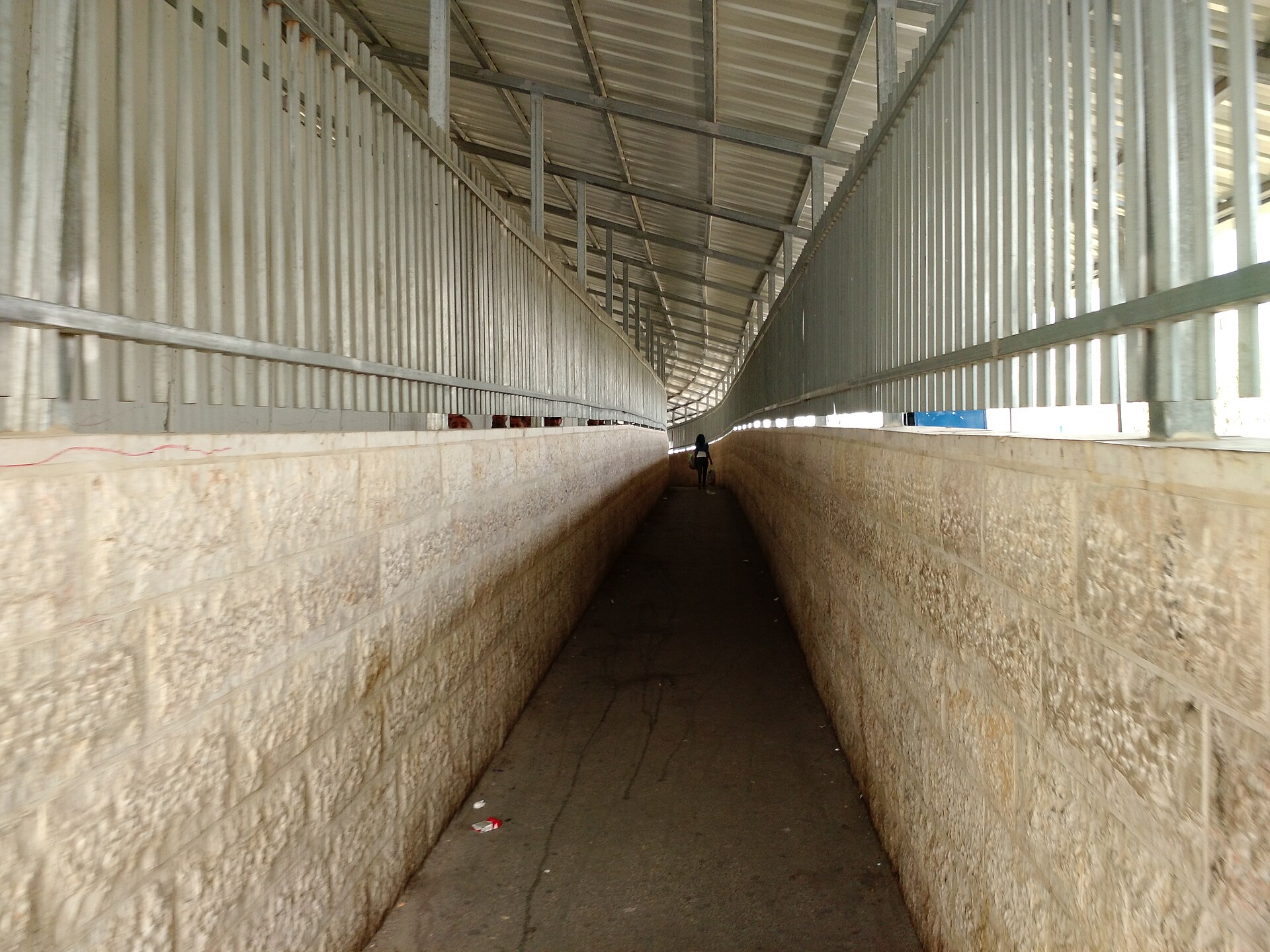 A long, narrow covered walkway between a stone wall and metal railings at Checkpoint 300 between Jerusalem and Bethlehem, with a single person walking in the distance