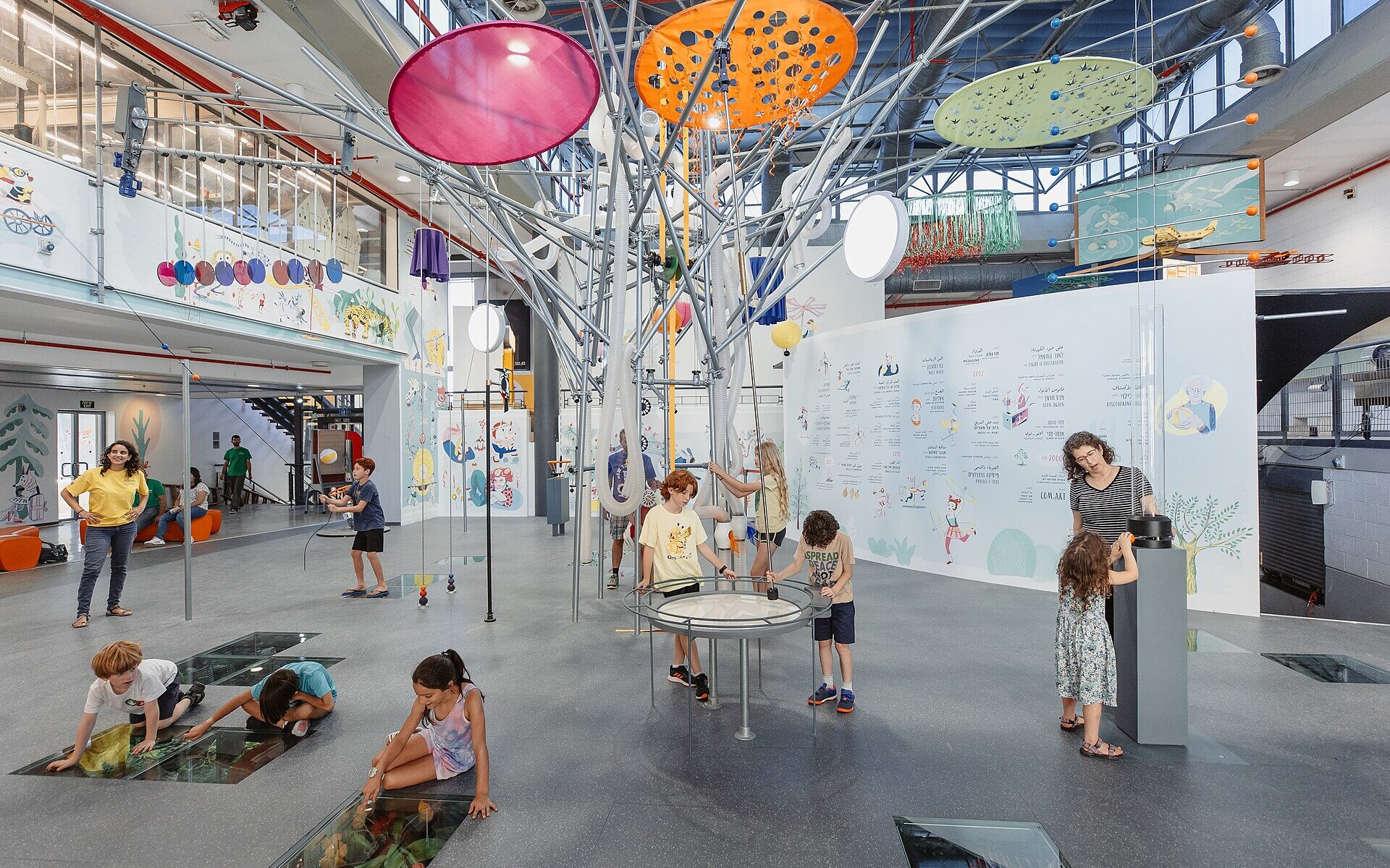 Children crouch over glass floor panels and gather around a round tabletop exhibit beneath colourful hanging discs in a bright hall of the Bloomfield Science Museum