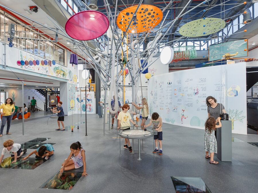 Children crouch over glass floor panels and gather around a round tabletop exhibit beneath colourful hanging discs in a bright hall of the Bloomfield Science Museum