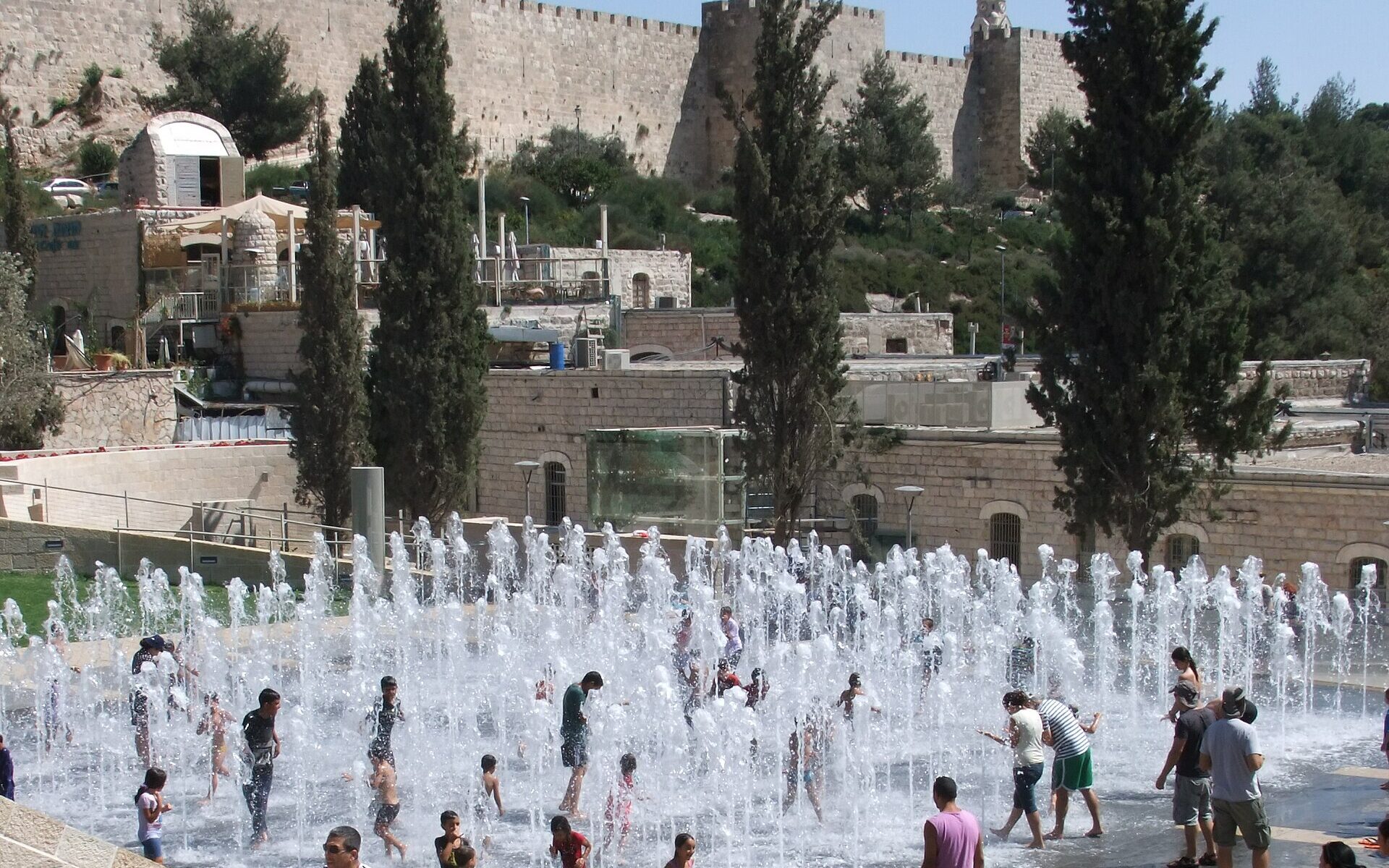 Children splash among dozens of water jets in the Teddy Park fountain, with parents looking on and the crenellated Old City walls and cypresses behind