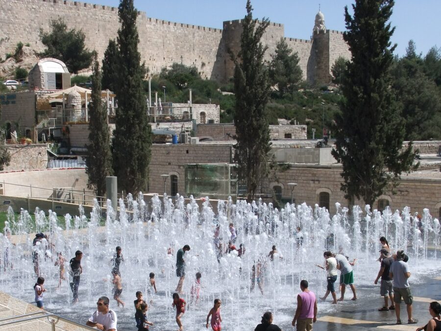 Children splash among dozens of water jets in the Teddy Park fountain, with parents looking on and the crenellated Old City walls and cypresses behind