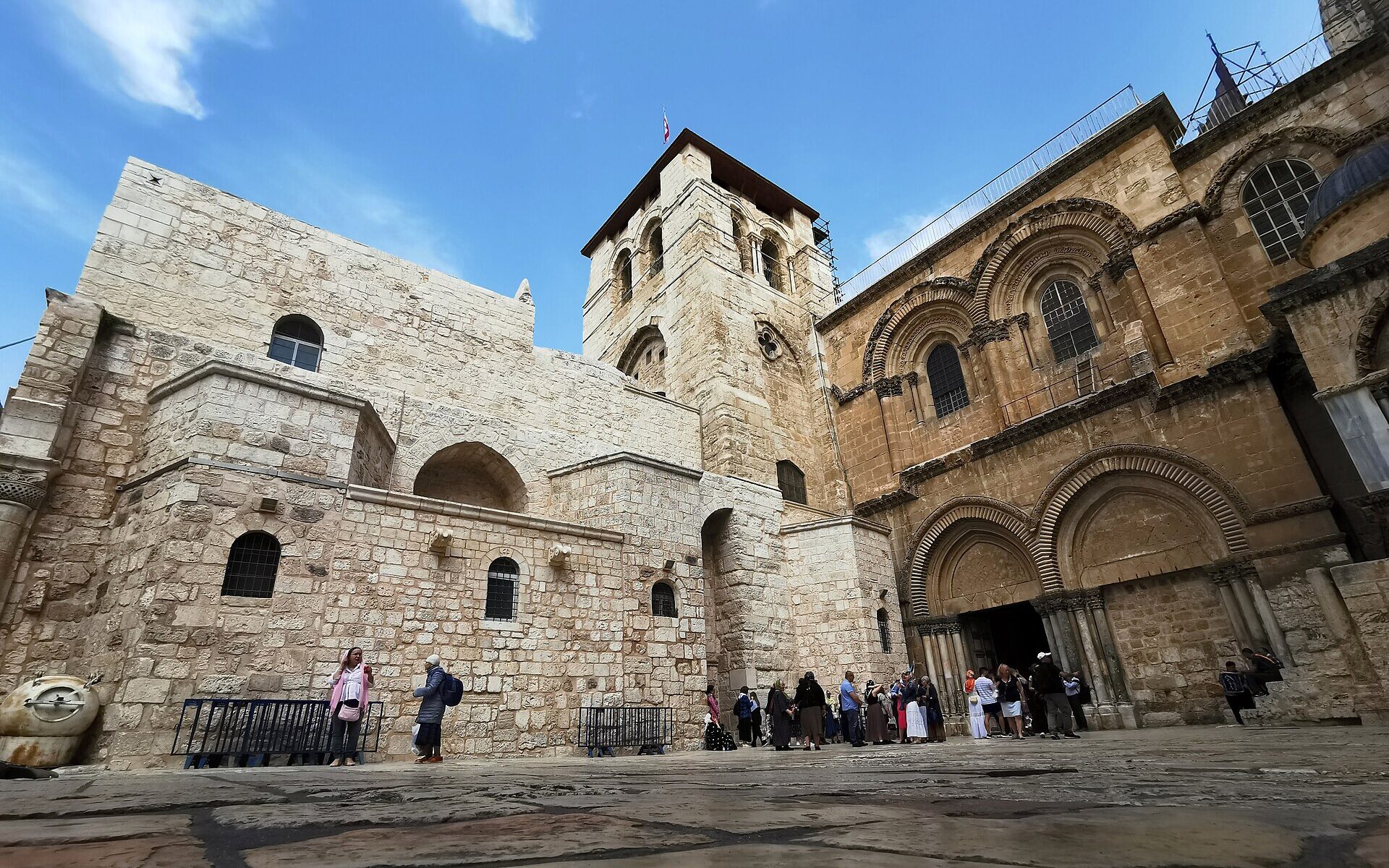 Church of the Holy Sepulchre seen from the parvis, with its square Crusader bell tower, twin-arched facade and open doorway, and visitors gathered at the entrance