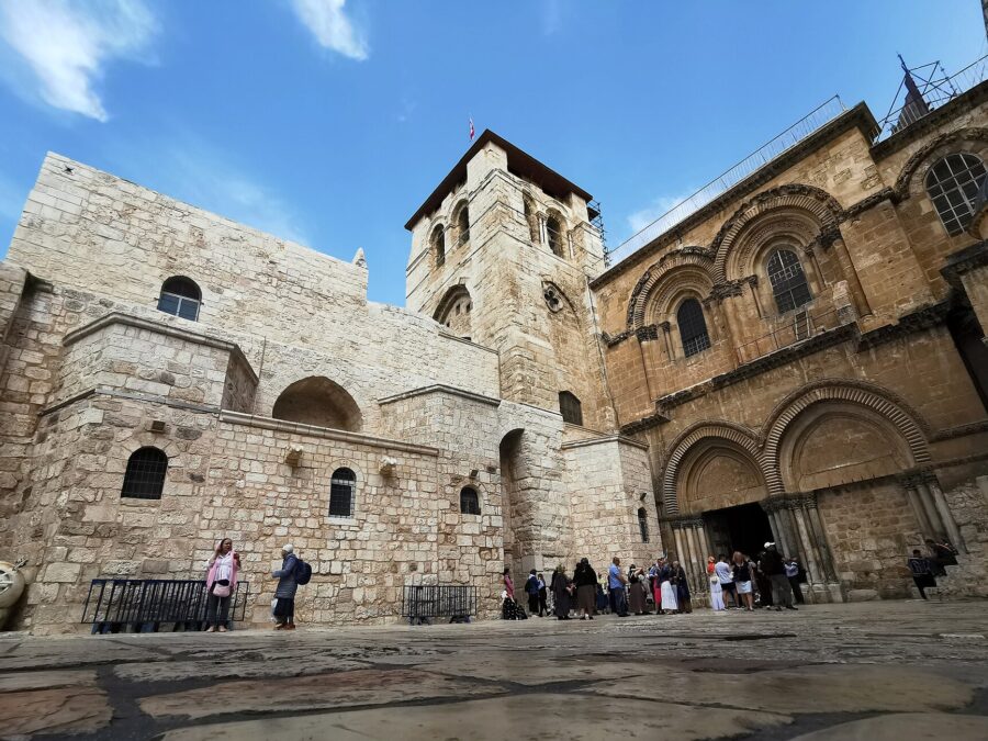 Church of the Holy Sepulchre seen from the parvis, with its square Crusader bell tower, twin-arched facade and open doorway, and visitors gathered at the entrance