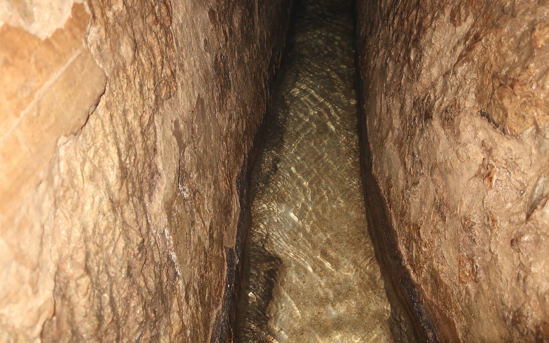Clear shallow water running along the narrow, rough-hewn limestone channel of Hezekiah's Tunnel, lit by a warm torch beam