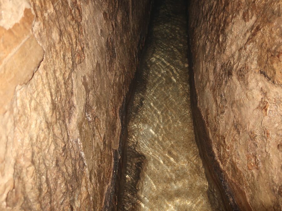 Clear shallow water running along the narrow, rough-hewn limestone channel of Hezekiah's Tunnel, lit by a warm torch beam