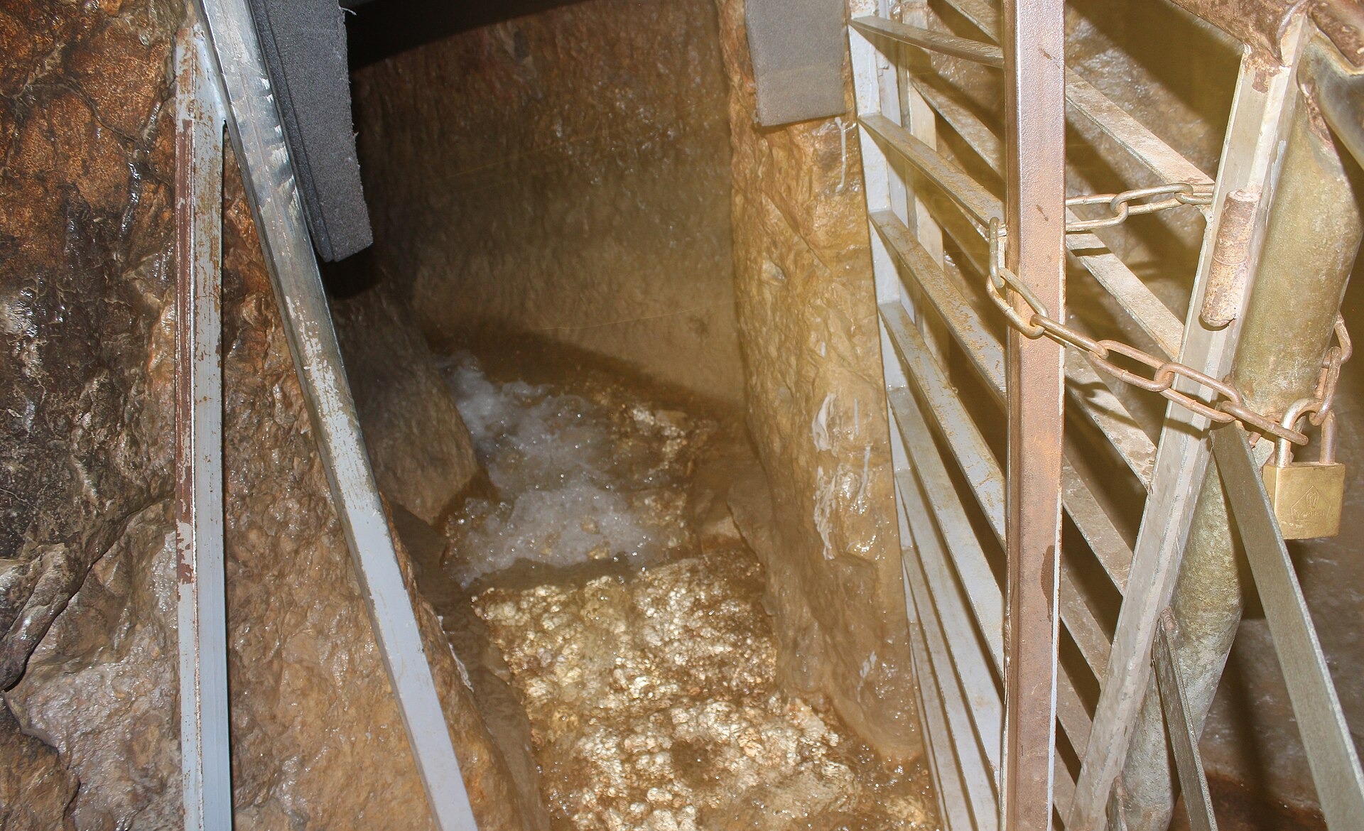 Clear spring water foaming over the rock floor of the narrow Gihon Spring cave, seen beside a metal gate secured with a chain and padlock