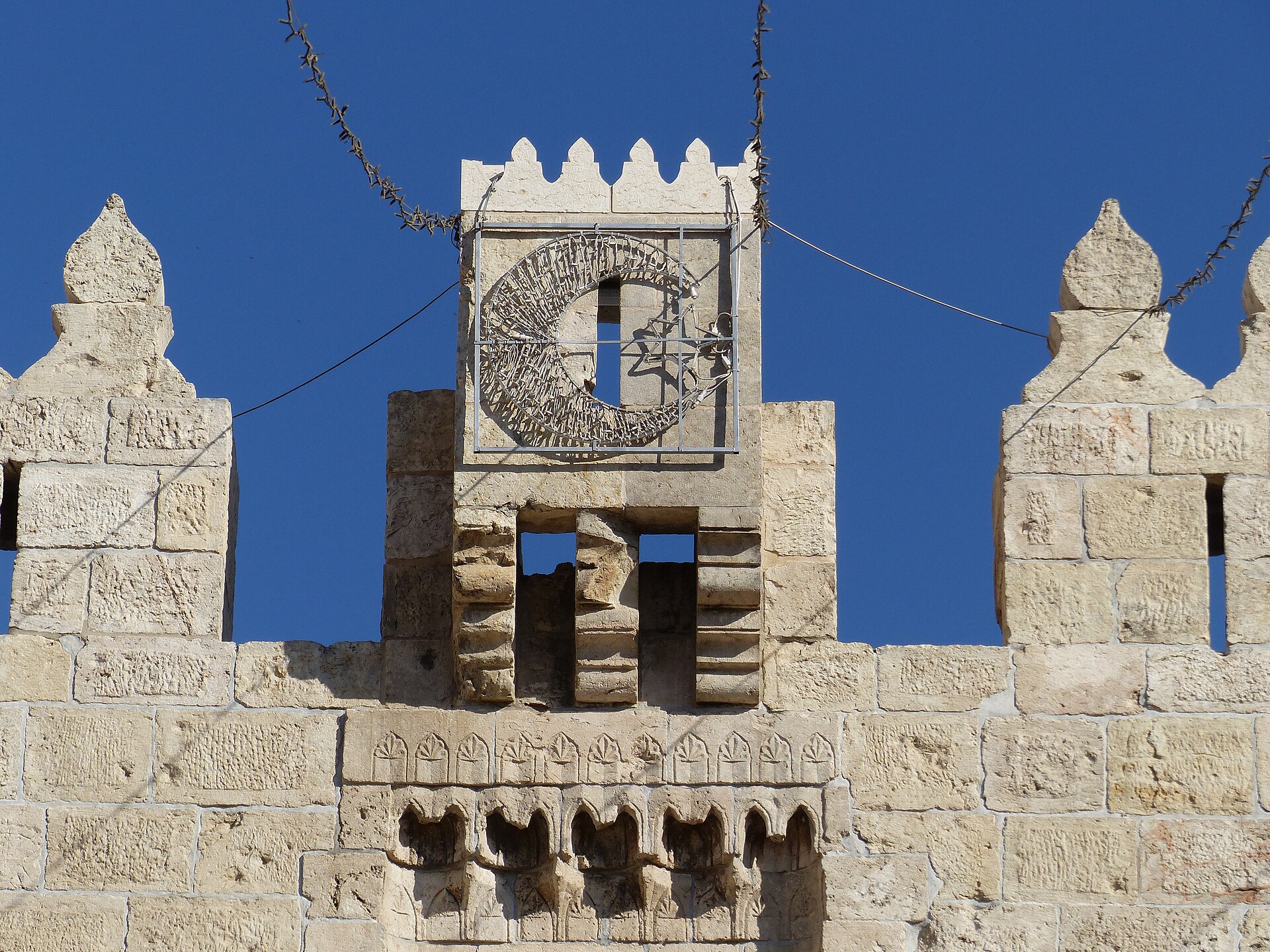 Close-up of the top of Damascus Gate: a small crenellated turret bearing a metal crescent frame, flanked by pointed stone merlons, above a carved frieze and corbels