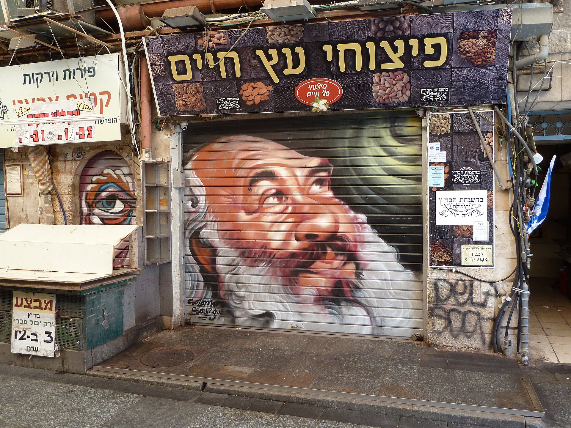 A closed shop shutter at Mahane Yehuda Market spray-painted by Solomon Souza with a large portrait of the singer Shlomo Carlebach, below a Hebrew nut shop sign