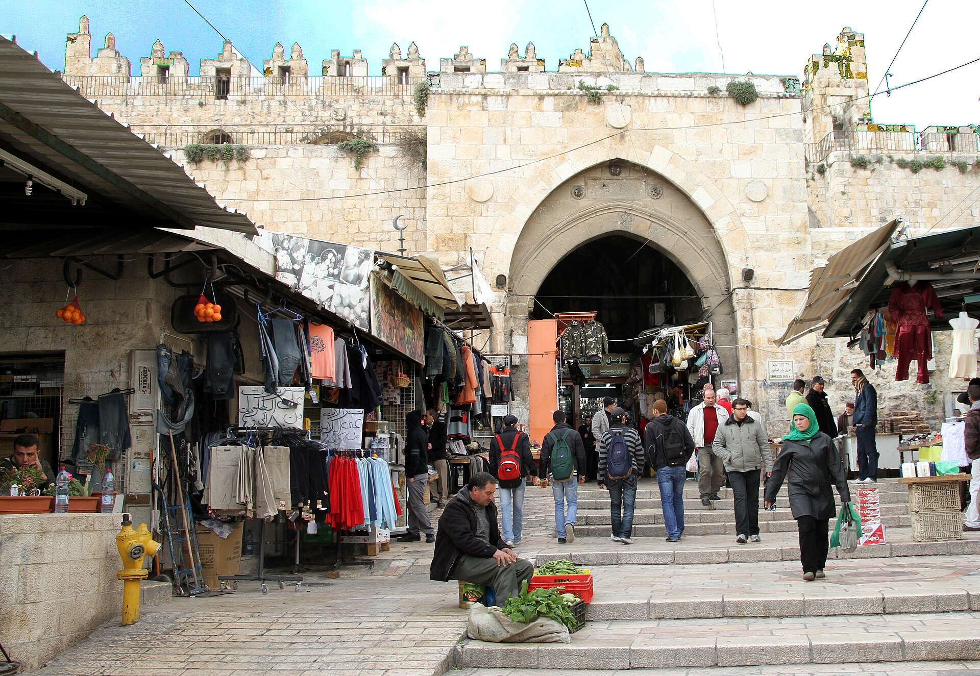 Clothes stalls and a vegetable seller on the stone steps inside Damascus Gate, with shoppers walking through the pointed arch of the gate's inner facade