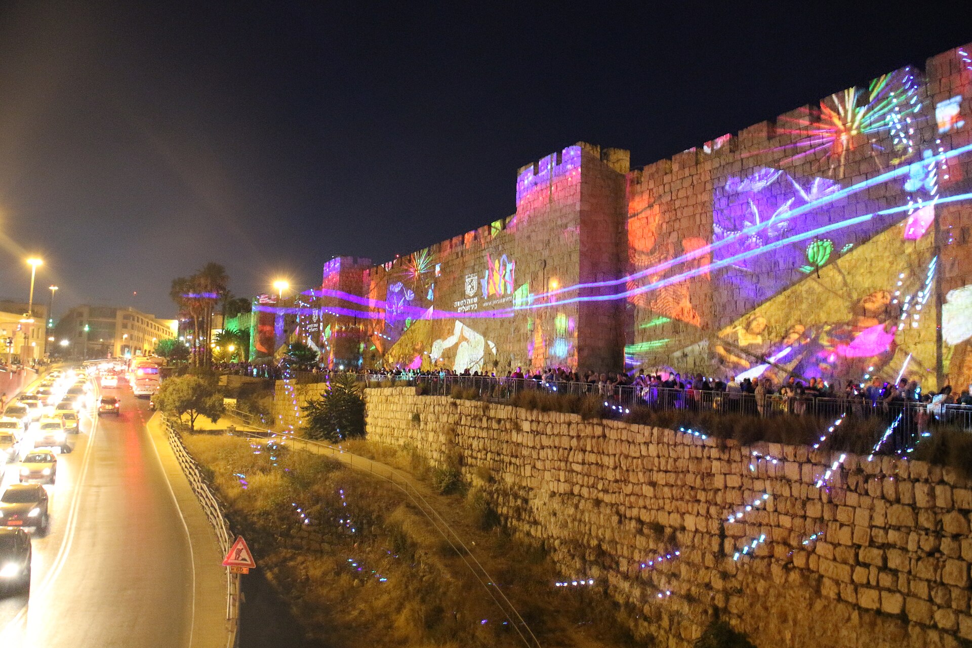 Colourful projections and laser lines covering the crenellated Old City wall near Jaffa Gate at night, with crowds along the path below and traffic on the road