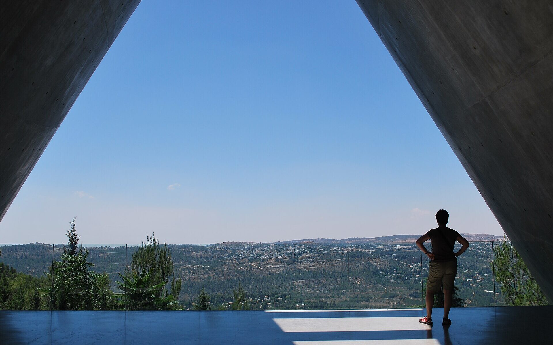 Concrete walls of Yad Vashem's Holocaust History Museum framing a wide view over forested Jerusalem hills, a lone visitor standing at the glass balcony rail