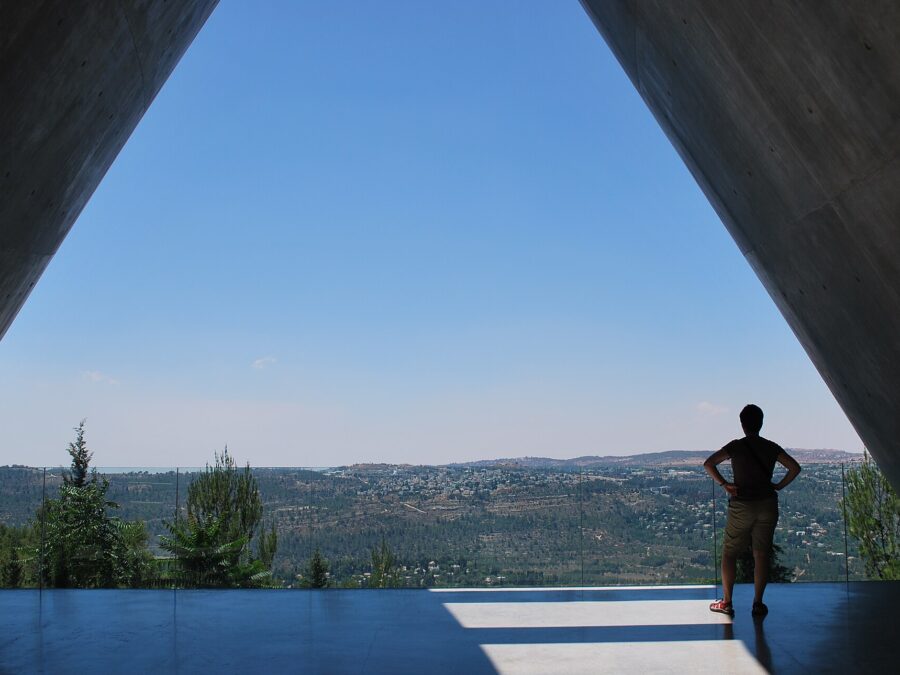 Concrete walls of Yad Vashem's Holocaust History Museum framing a wide view over forested Jerusalem hills, a lone visitor standing at the glass balcony rail