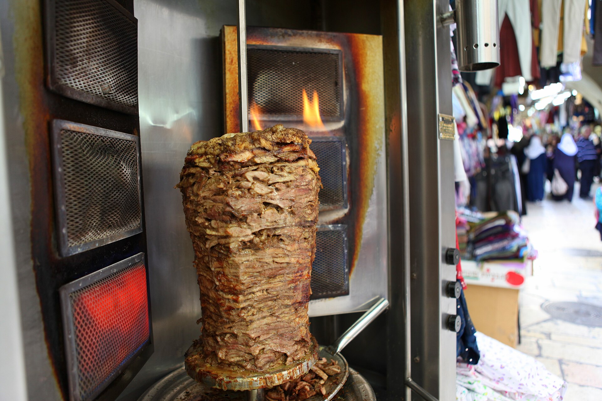 Cone of layered shawarma meat roasting on a vertical spit beside a flaming grill, with a busy Old City market alley blurred in the background