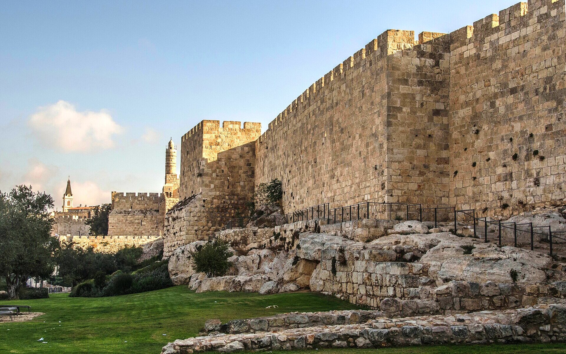 The crenellated Ottoman wall of Jerusalem's Old City rising from bedrock in early-morning sun, with a square tower and the Tower of David minaret in the distance
