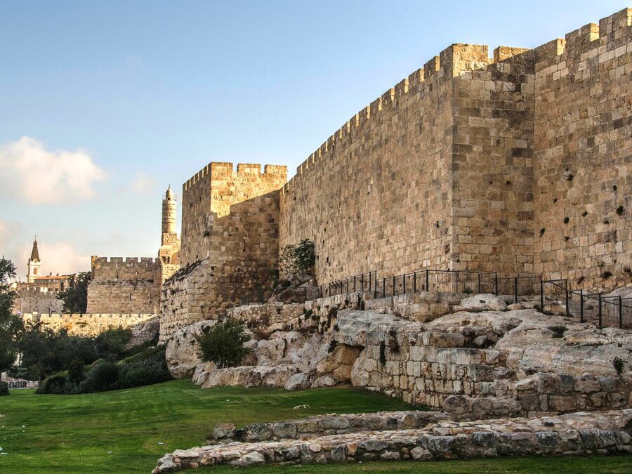 The crenellated Ottoman wall of Jerusalem's Old City rising from bedrock in early-morning sun, with a square tower and the Tower of David minaret in the distance